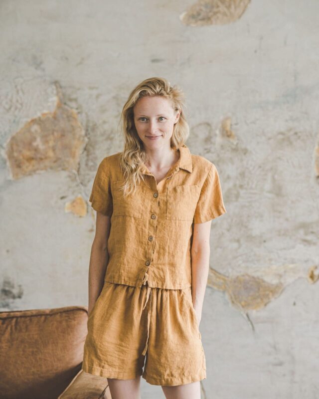 Ochre linen shirt and matching shorts by Old Linen Mill, worn against a bare plaster wall.